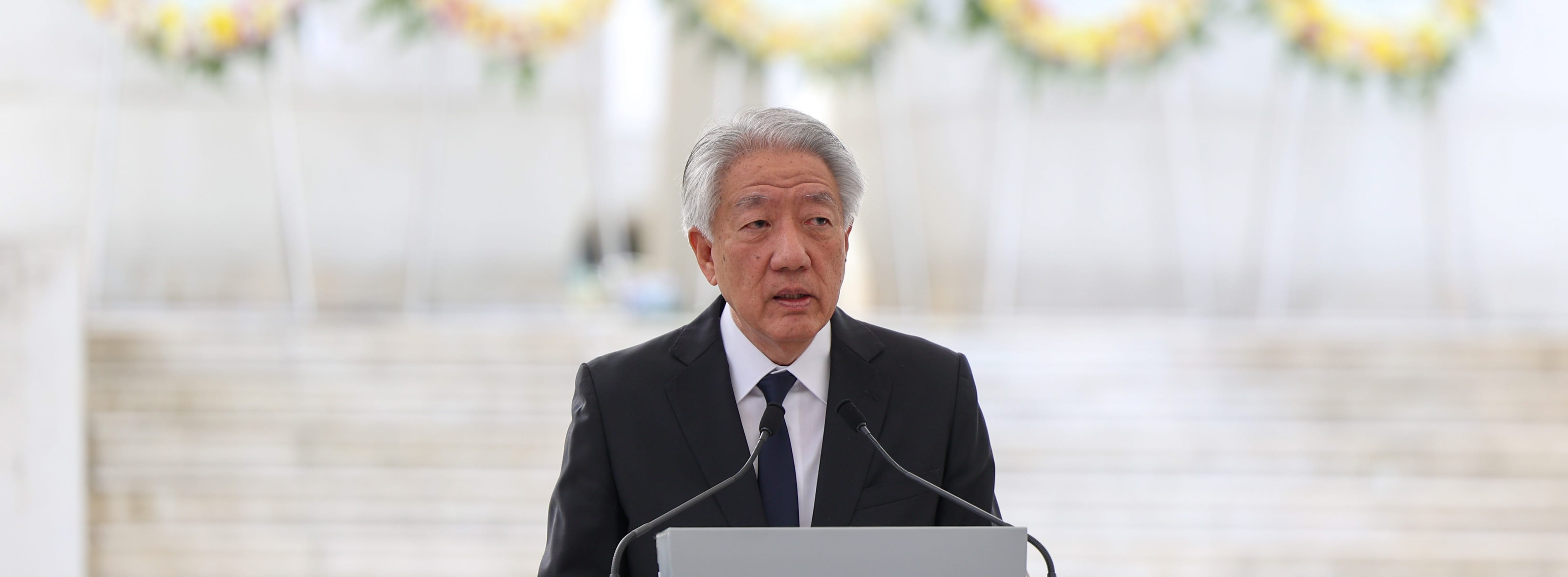 Man in a suit at podium speaking; floral wreaths in background.